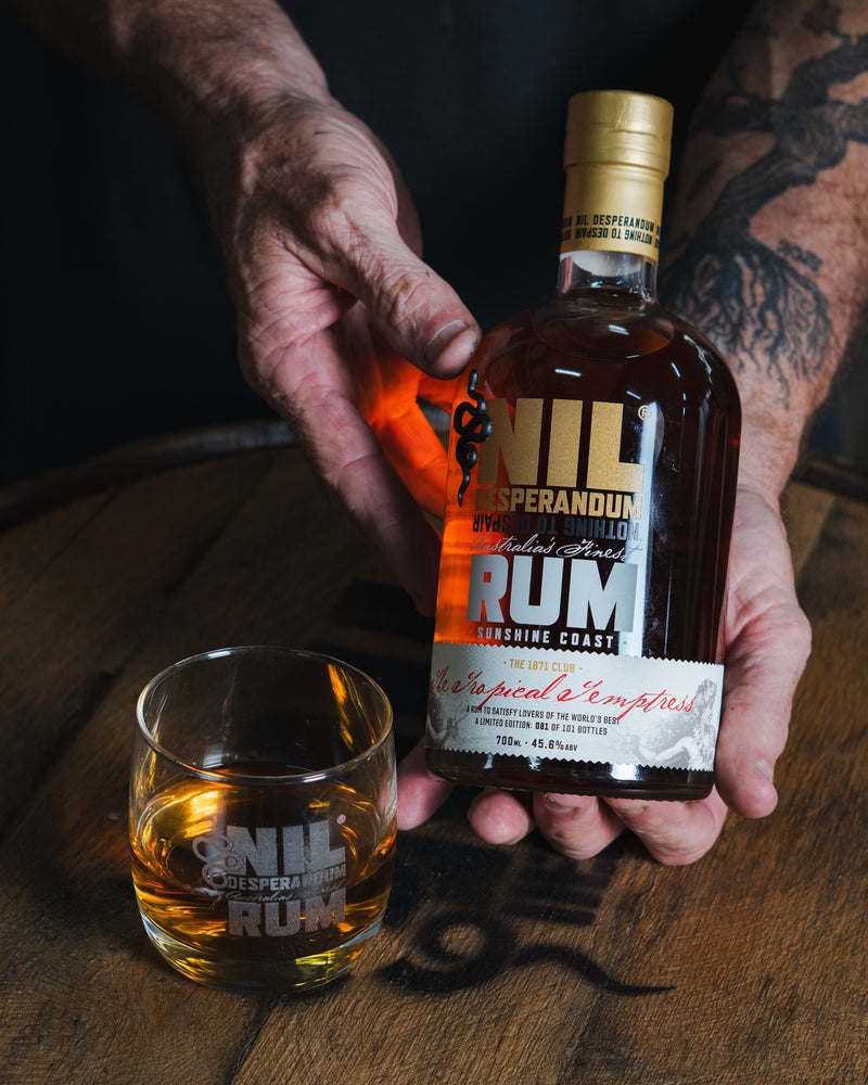 Hand holding a bottle of Nil Desperandum Rum with a glass on a wooden surface