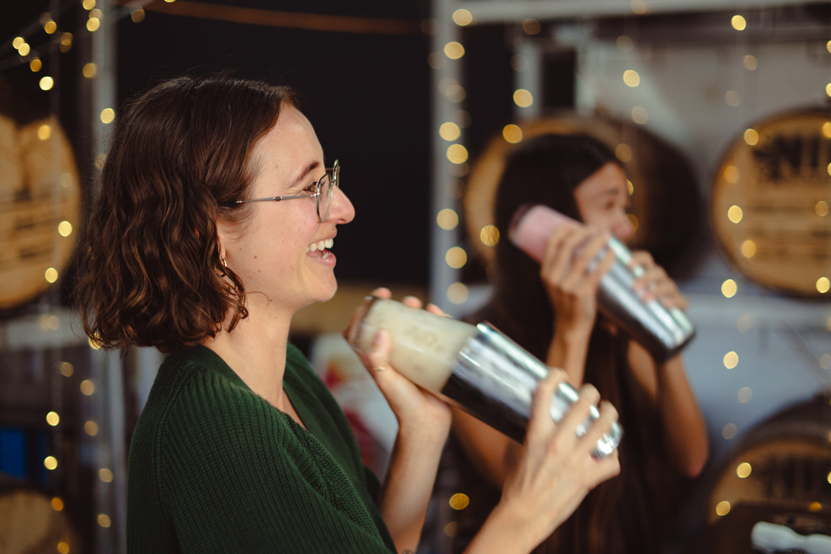 Two women in a warmly lit indoor setting, each holding a stainless steel cocktail shaker with a drink inside. The foreground woman wears a dark green top and glasses, while the background woman wears a pink top.